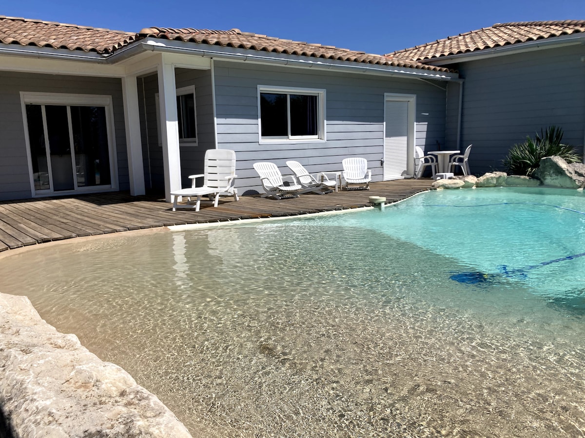 A serene lagoon-style pool is framed by smooth rock edges, with clear water reflecting sunlight. Several white lounge chairs are positioned on a wooden deck nearby, adjacent to a light-colored house with expansive glass doors.