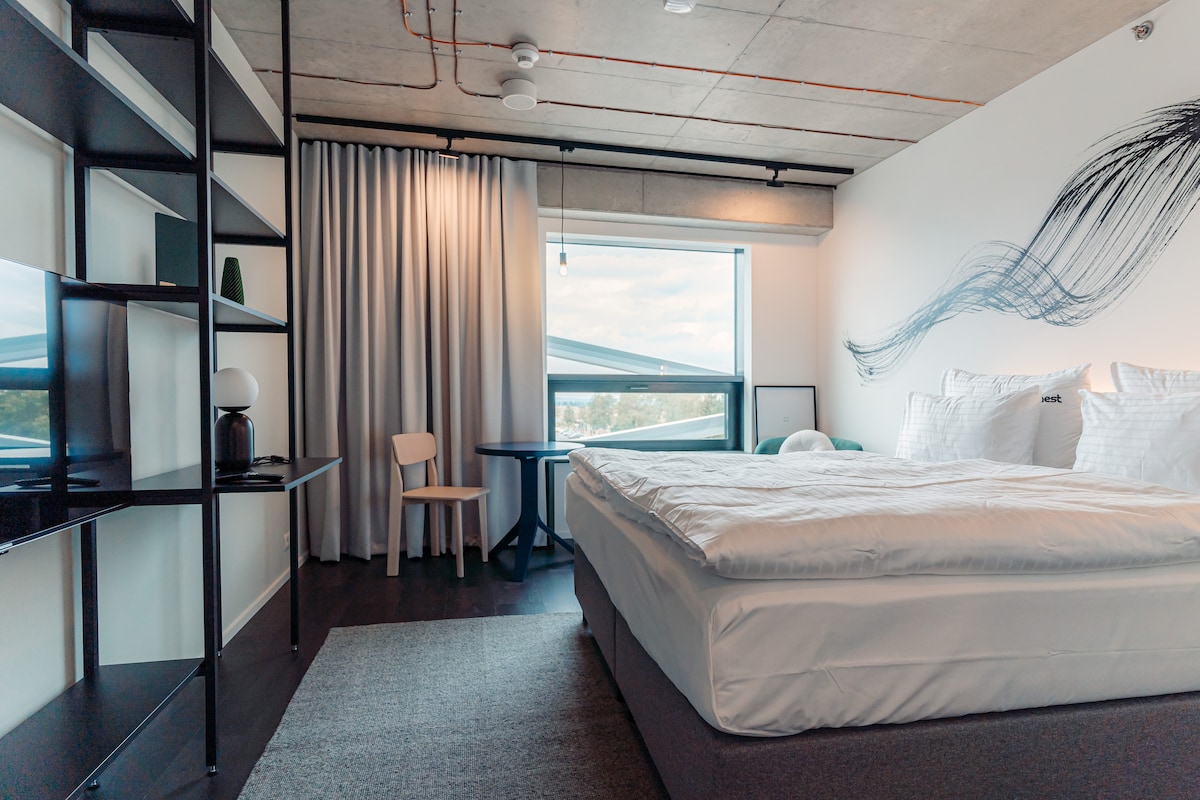 A modern bedroom features a spacious bed with crisp linens, complemented by a minimalist bookshelf and a light wooden chair. A small table sits nearby, positioned under a large window that allows natural light to fill the room, framed by soft drapes.