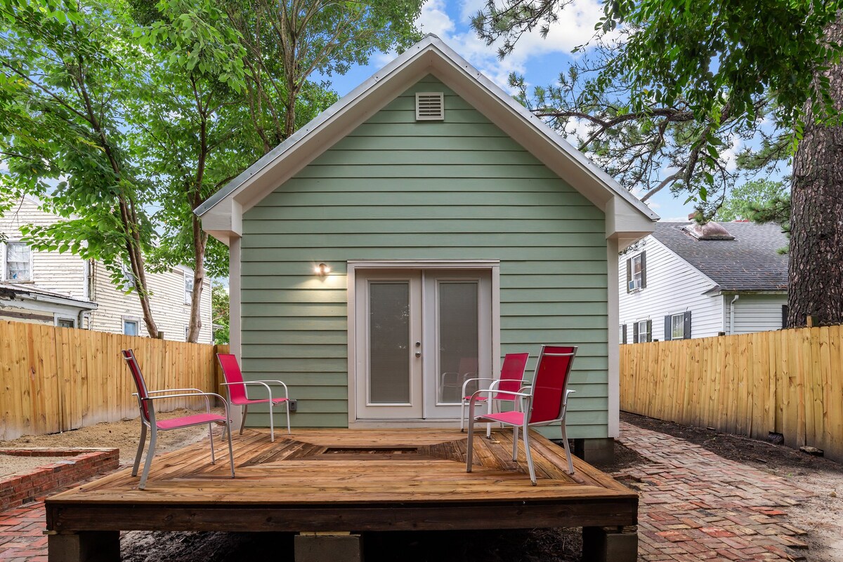 A newly renovated cottage is displayed from the rear, showcasing a wooden deck with four pink chairs. The green exterior of the cottage is complemented by surrounding trees, and a fenced yard provides privacy. The entrance features sliding glass doors allowing for natural light.