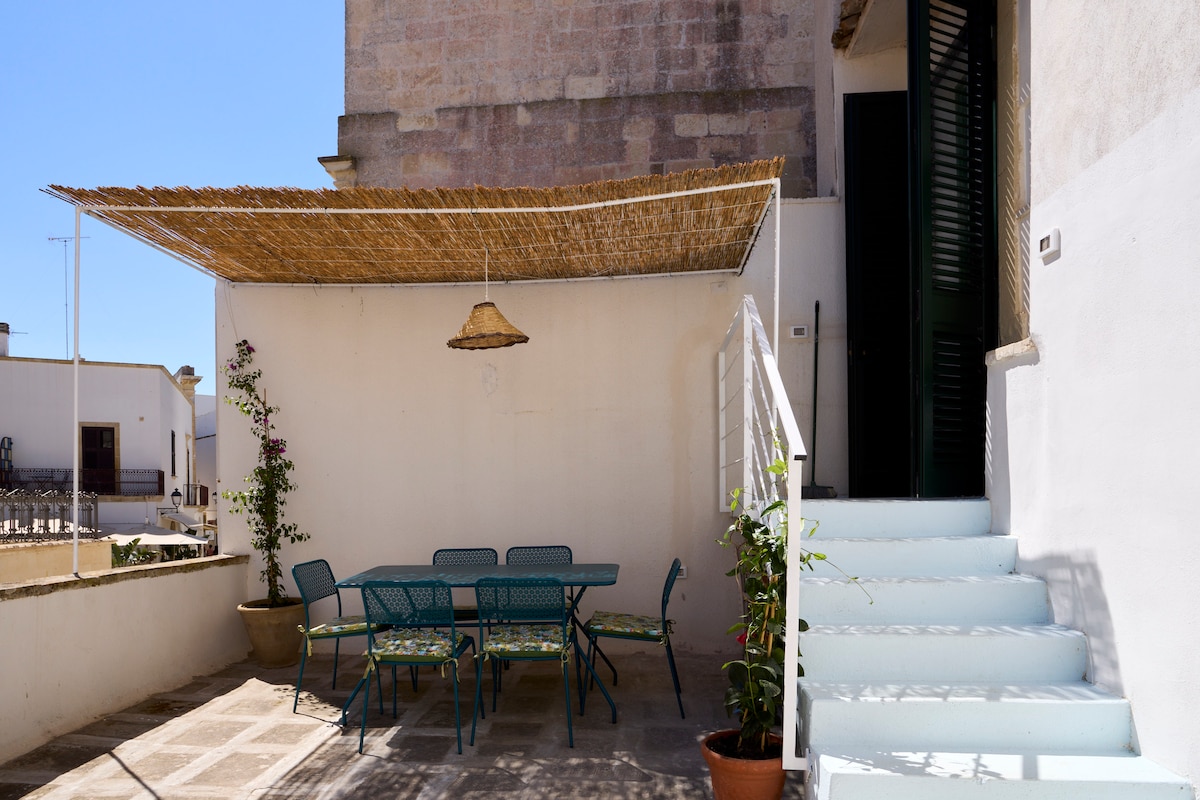 An outdoor terrace is presented with a shaded area featuring a straw umbrella. A rectangular table with multiple chairs is set up, surrounded by potted greenery. White stairs lead up to an entrance, complemented by a plain wall backdrop.
