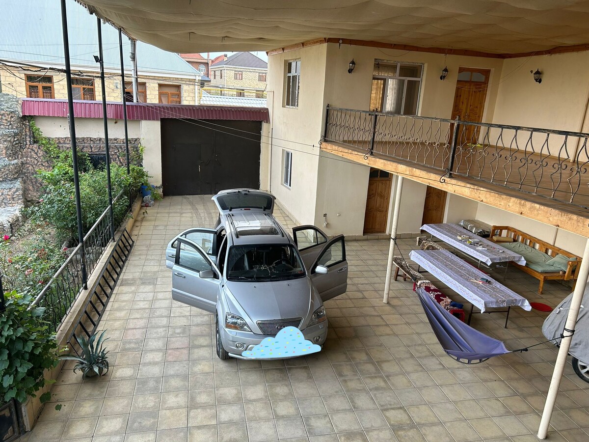 An open-air courtyard features a silver car parked with its doors ajar, surrounded by a tiled floor. Several outdoor seating areas are evident, including a wooden bench and tables adorned with light-colored tablecloths. Green plants in pots add a touch of nature to the space.