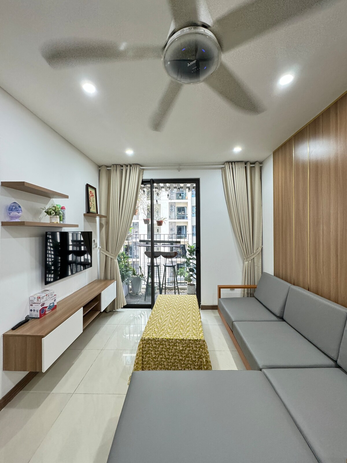 A modern living area is presented with a sleek gray sofa and a light wood accent wall. A flat-screen television is mounted on the wall, and natural light floods in through glass doors leading to a balcony. Light-colored curtains frame the windows, adding softness to the space.