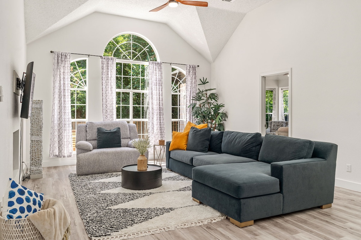 A bright living area features a spacious dark gray sectional sofa positioned on a patterned rug. Large windows allow natural light to fill the room, with sheer curtains gently framing the view. A decorative plant and a cozy armchair add inviting touches to the space.