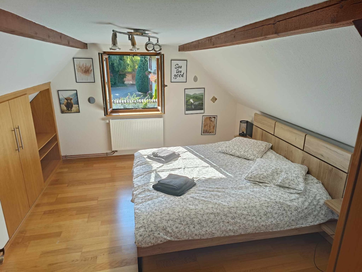 An inviting bedroom featuring a spacious bed adorned with soft linens, complemented by decorative wall art. Natural light filters through a window, illuminating the wooden flooring and simple shelving unit. A view of greenery outside enhances the serene atmosphere.