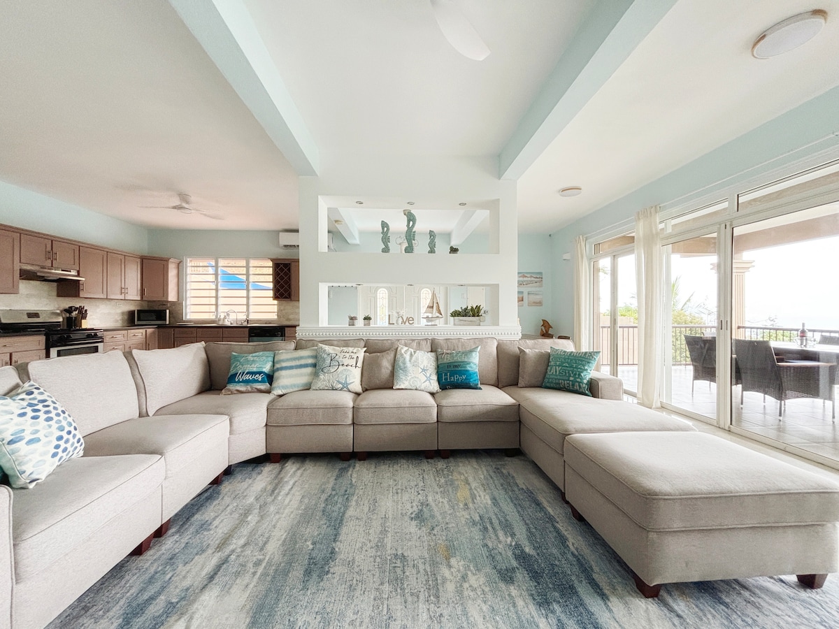 A spacious living area featuring a large sectional sofa adorned with decorative pillows. Natural light floods the room through expansive sliding glass doors leading to the terrace. A soft area rug covers the floor, complementing the light-colored walls and ceiling.