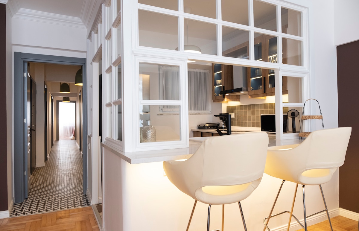The kitchen features a half-wall design with large windows, providing a view of the space. Two high-backed stools are positioned at a sleek counter. Soft lighting illuminates the area, complementing the modern cabinetry and tiled backsplash. A hallway extends in the background, leading to the rest of the apartment.