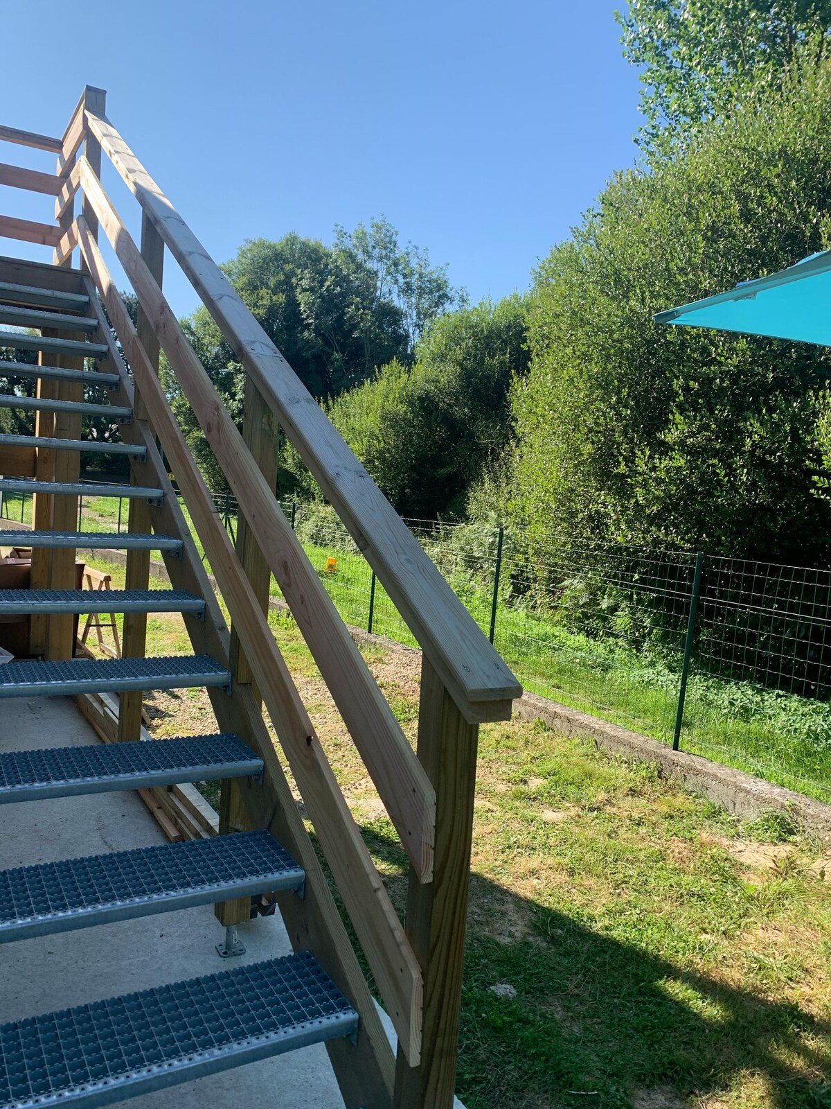 An outdoor staircase made of wood and metal leads to an upper level, surrounded by greenery. A bright blue umbrella is visible nearby, providing shade over an outdoor area. The background features lush trees and a gentle slope of grass under a clear blue sky.