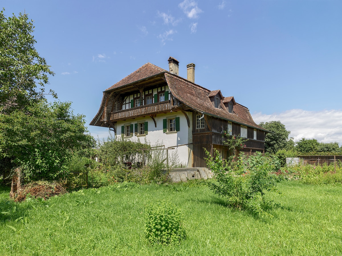 A charming historic building is set amidst lush green grass, showcasing a traditional wooden structure with a sloped roof and multiple balconies. The exterior features decorative window shutters, and the surrounding greenery enhances the tranquil atmosphere.