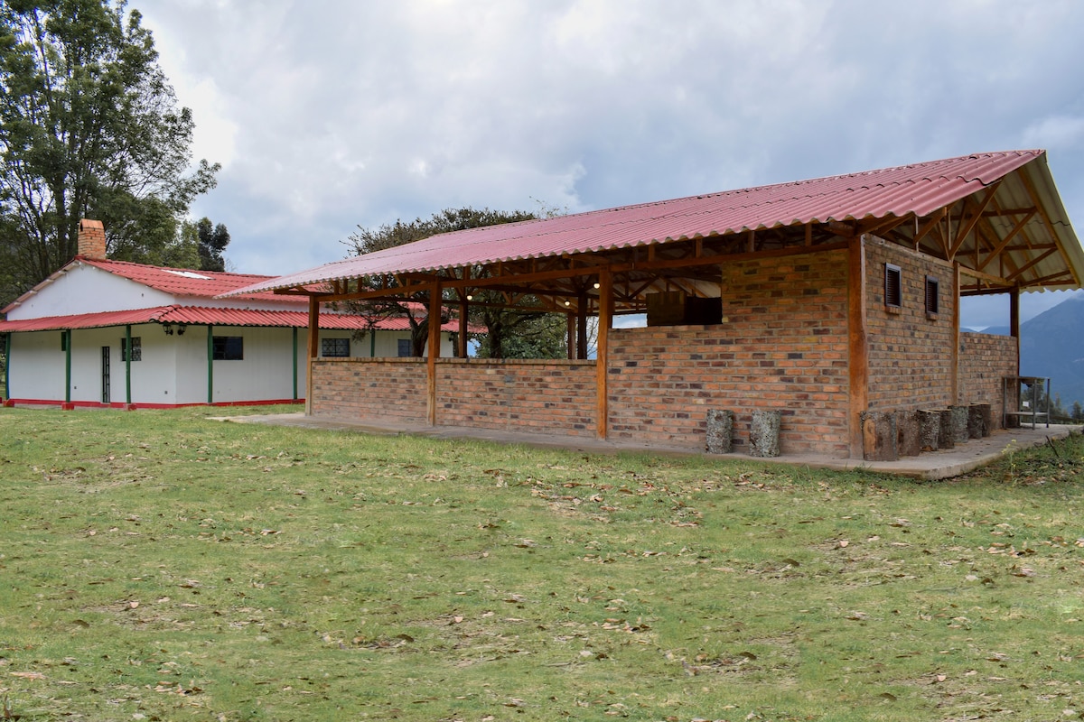 A charming country house showcases rustic architecture with a red-tiled roof and stone accents. Surrounding green lawns create an inviting atmosphere. Several outdoor seating arrangements are visible under the extended roof, offering a shaded area for relaxation or gatherings.