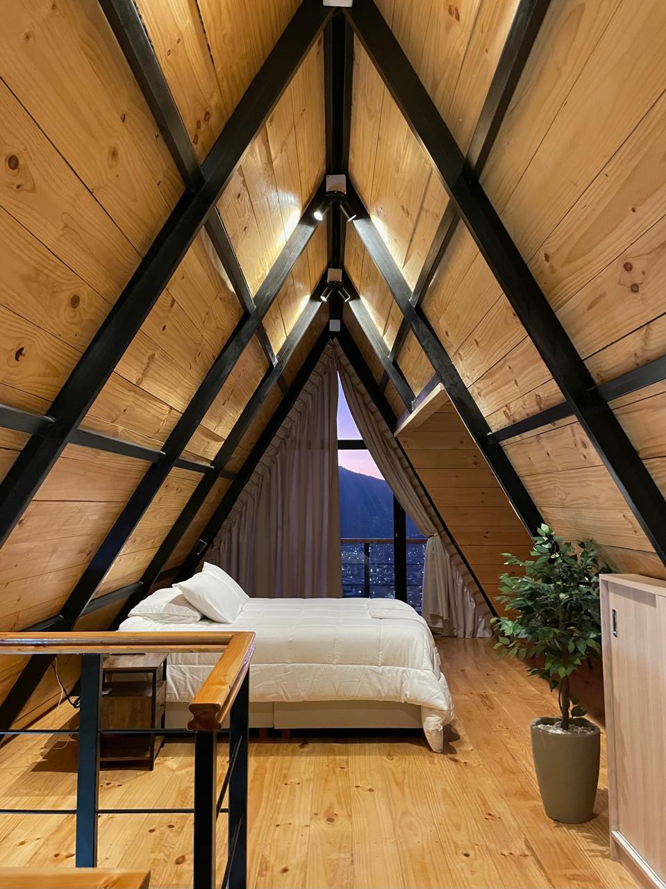 A spacious bedroom features an A-frame design with wooden walls and beams. A comfortable bed is positioned against a large window offering views of the mountains at dusk. A small plant adds a touch of greenery, enhancing the serene atmosphere.