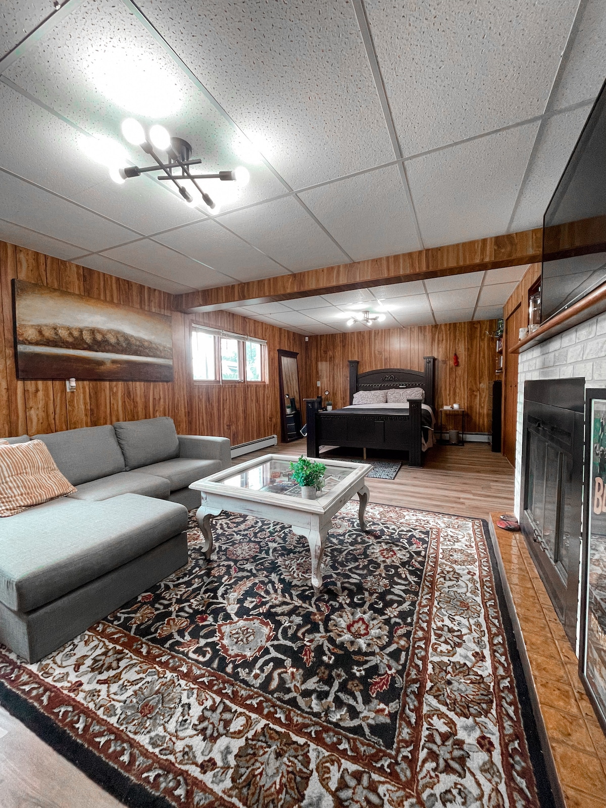 An open concept living space features a sectional couch and a coffee table on a patterned area rug. A spacious bed is positioned in the background, with natural light filtering through two windows. A fireplace adds warmth to the inviting atmosphere.
