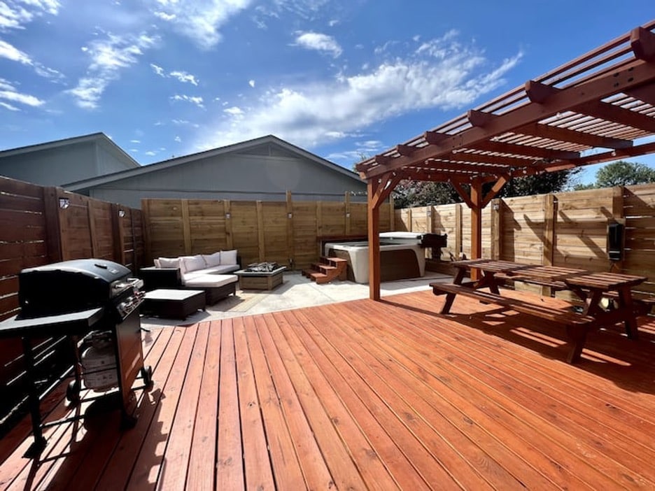 A spacious outdoor deck features a shaded pergola, a hot tub, and seating arrangements, including a cozy sectional and picnic-style table. An outdoor grill is positioned nearby, while wooden fencing provides privacy and a sense of seclusion in the stylish backyard.