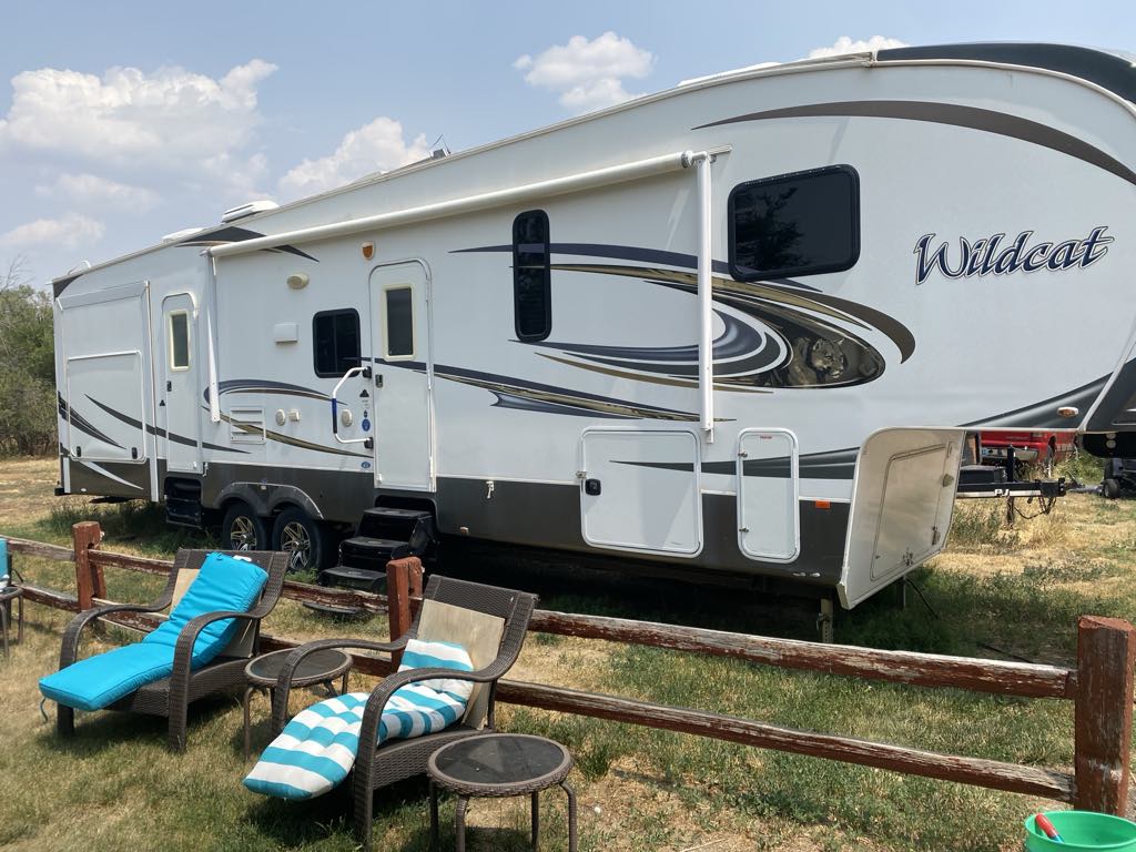 A spacious RV is positioned on a grassy area, surrounded by a wooden fence. Two lounge chairs with striped cushions are placed nearby, offering a relaxing spot under a clear sky. The RV features large windows and a sleek design, enhancing its modern appearance.