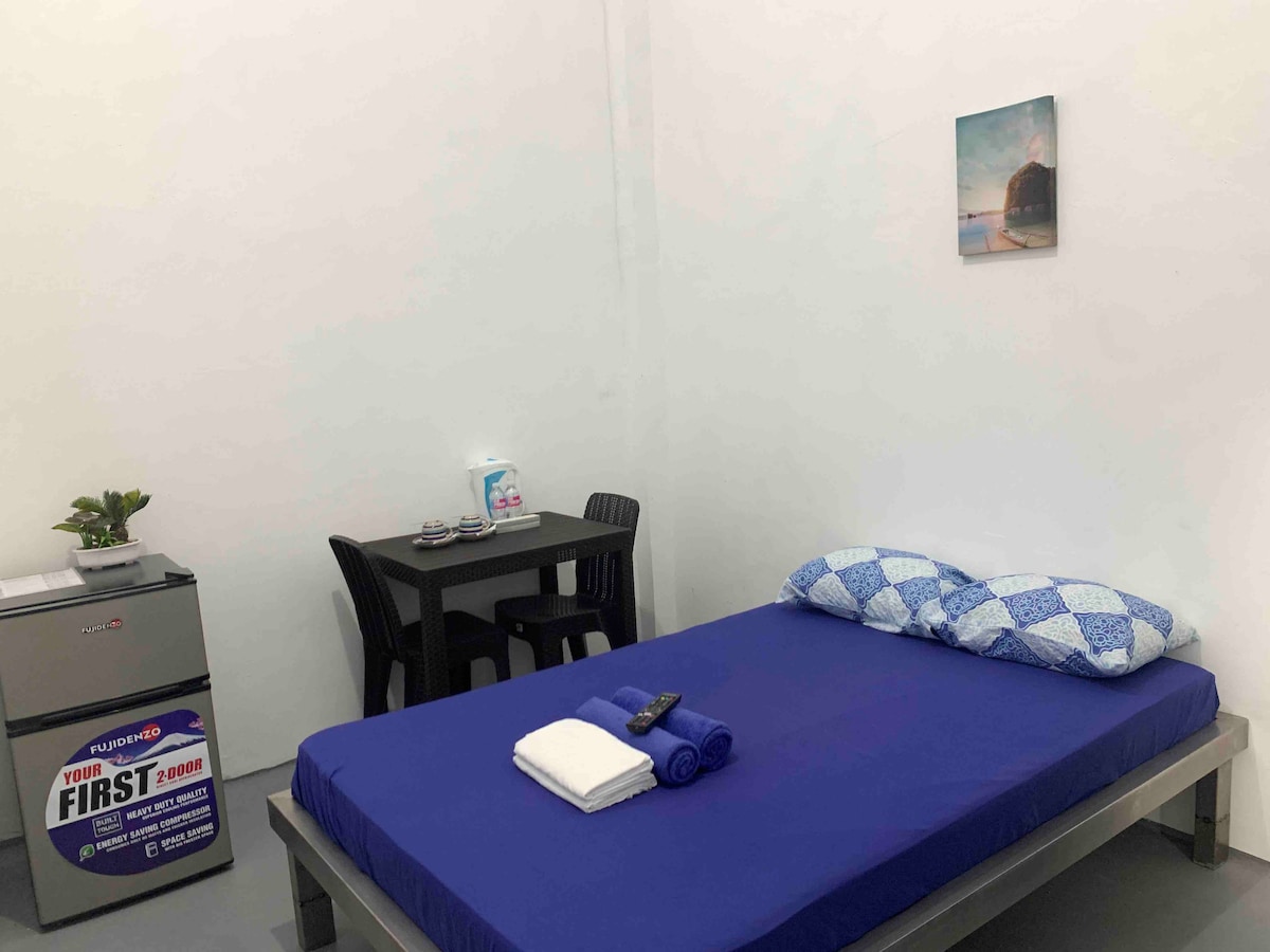 A cozy sleeping area features a spacious bed dressed in blue linens. A small table with four chairs is nearby, alongside a mini-fridge. A painting of a beach scene adorns the wall, and clean towels are neatly arranged on the bed.