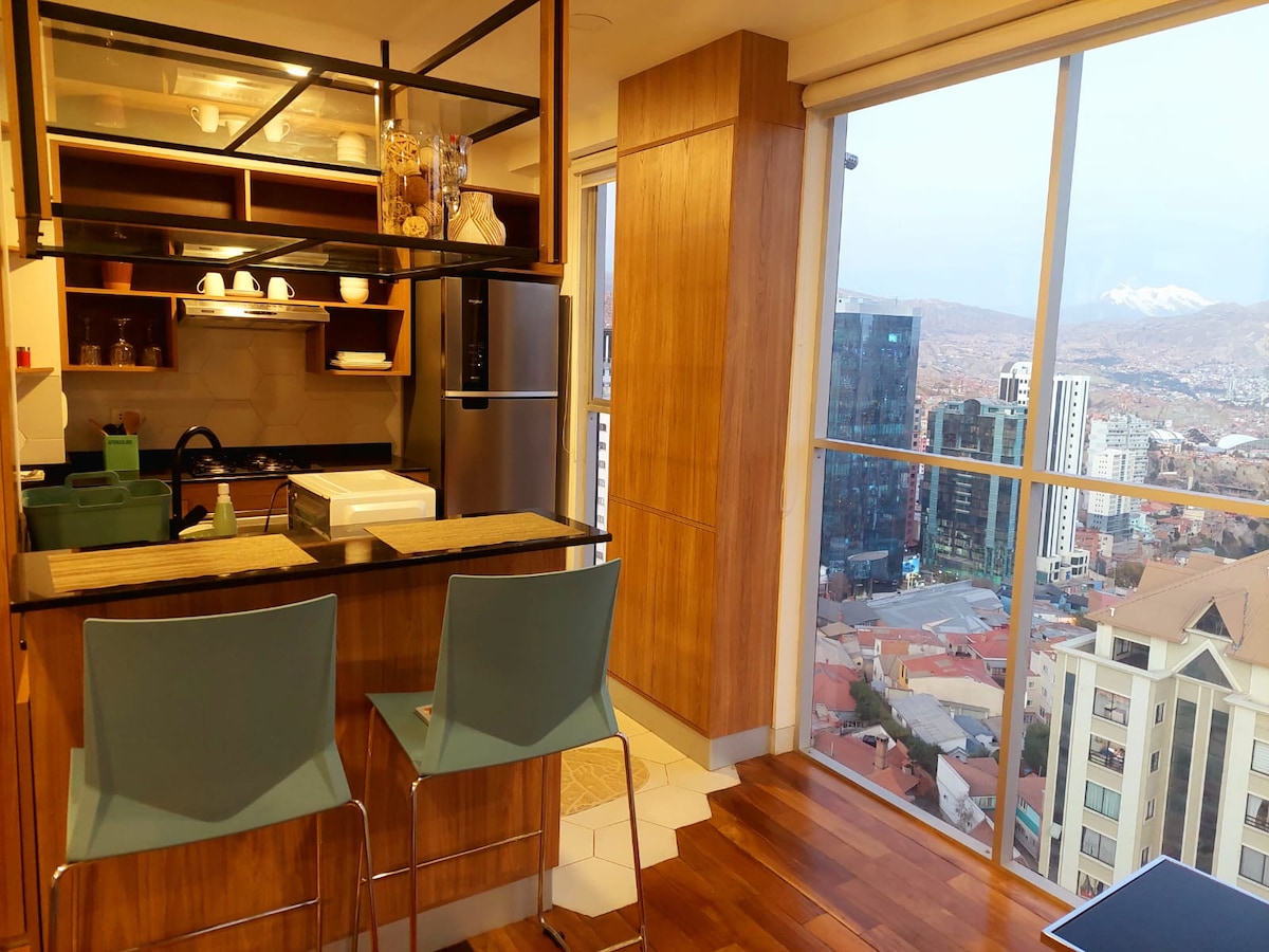 A modern kitchen area is visible, featuring sleek wooden cabinetry and a breakfast bar with two stools. Large floor-to-ceiling windows offer a panoramic city view, with a snow-capped mountain in the distance. Natural light brightens the open space, complemented by a subtle color palette.