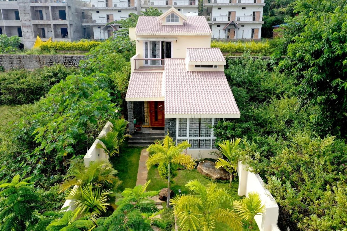 The villa is surrounded by lush greenery, featuring a well-maintained garden with various plants. The exterior showcases a light-colored structure with a pink tiled roof and large windows. A pathway leads to the entrance, framed by vibrant foliage.