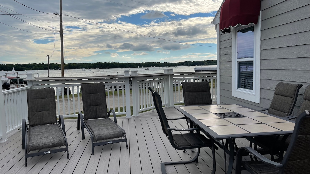 A spacious deck is furnished with a dining table and several comfortable lounge chairs. A scenic view of Lake Beulah is visible in the background, showcasing calm waters and a cloudy sky. The deck features a wooden railing and is partially shaded by an overhang.