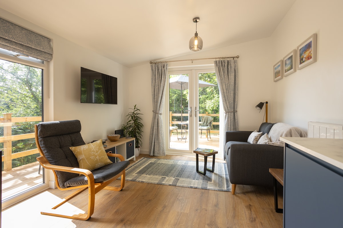 A cozy living area features a comfortable armchair alongside a sectional sofa. Natural light fills the space through glass doors leading to a deck, with outdoor seating visible. A decorative rug covers the wooden floor, and framed artwork is displayed on the wall.
