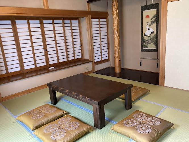 The tatami-floored room features a low wooden table at its center, surrounded by decorative cushions. Sliding shoji doors allow for soft natural light, while a wall-mounted artwork depicting a crane adds a cultural touch to the serene space.