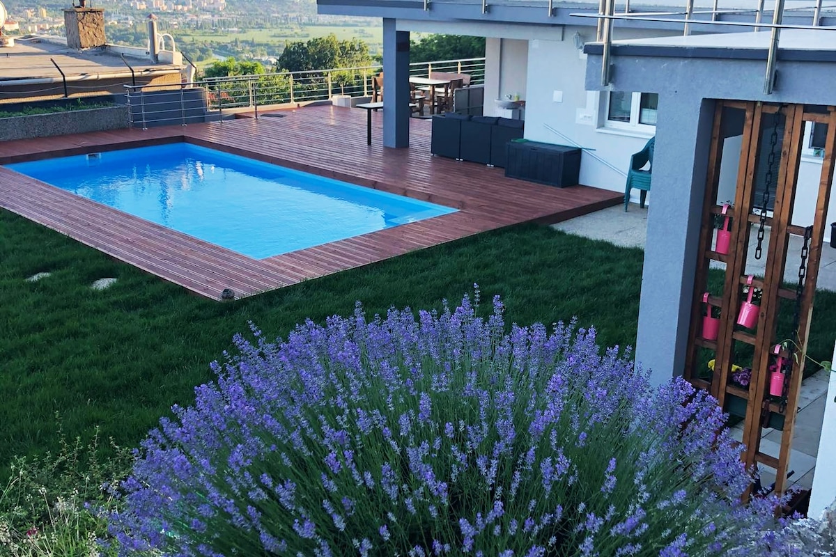 An outdoor space features a swimming pool surrounded by a wooden deck and a well-maintained grassy area. Lush lavender plants are visible in the foreground, adding a touch of greenery. Comfortable seating arrangements are present near the pool, with views of the surrounding landscape.