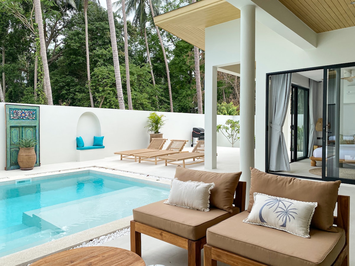 An outdoor living area is visible, featuring two wooden armchairs with light cushions positioned near a private salt-water pool. Sun loungers are arranged around the pool, with lush greenery providing a tranquil backdrop. Large sliding glass doors lead to a bright indoor space.