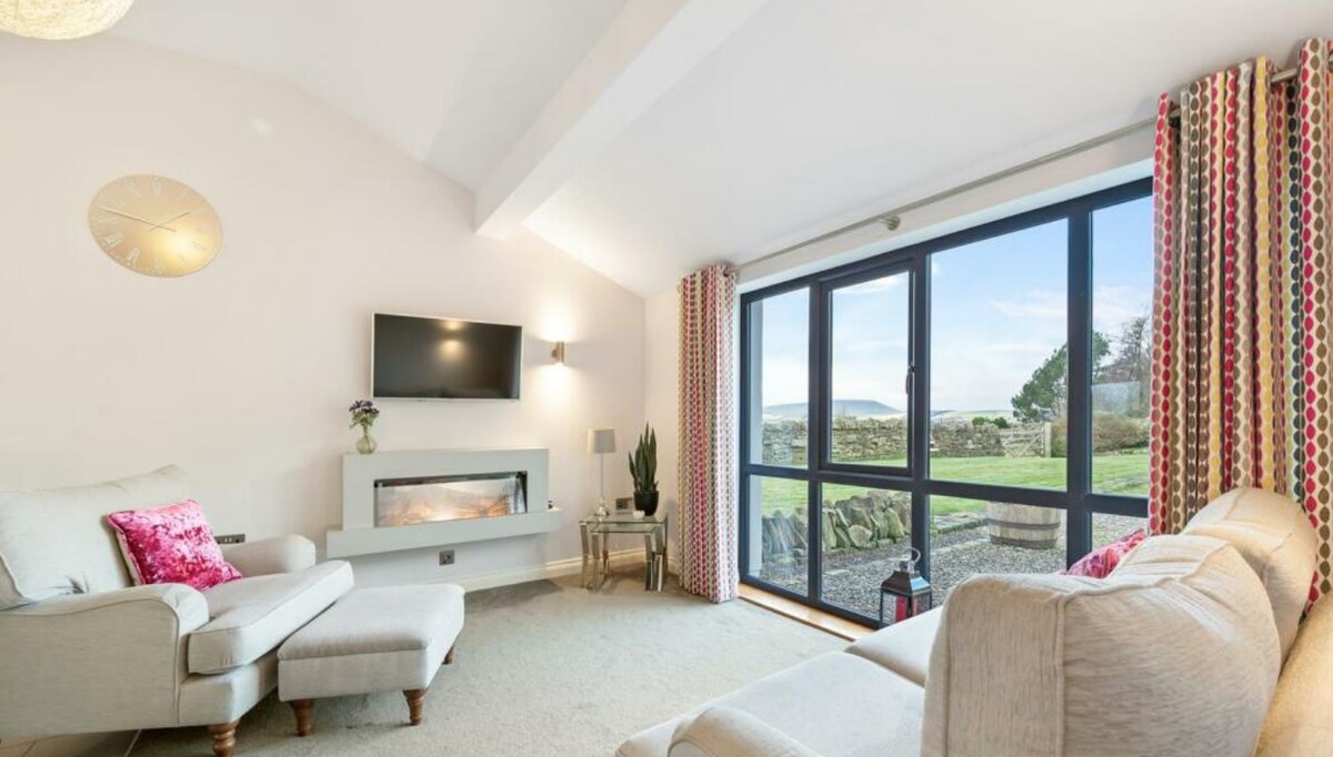 A light-filled living area features large glass doors that open to scenic views. Two comfortable armchairs are positioned invitingly, with a modern fireplace as a focal point. Soft, neutral colors complement patterned curtains, creating an airy and relaxed environment.