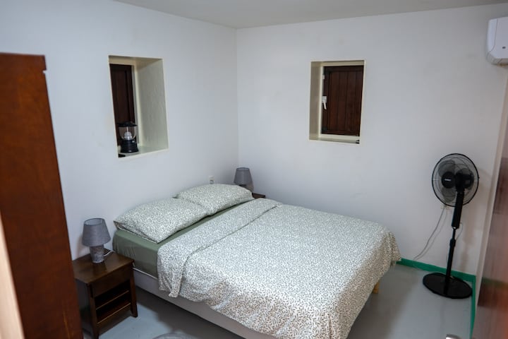 这间卧室配有两张床头柜，配有床头灯、空调、应急灯、衣柜、立式风扇和一张带半正面床垫的弹簧床。 