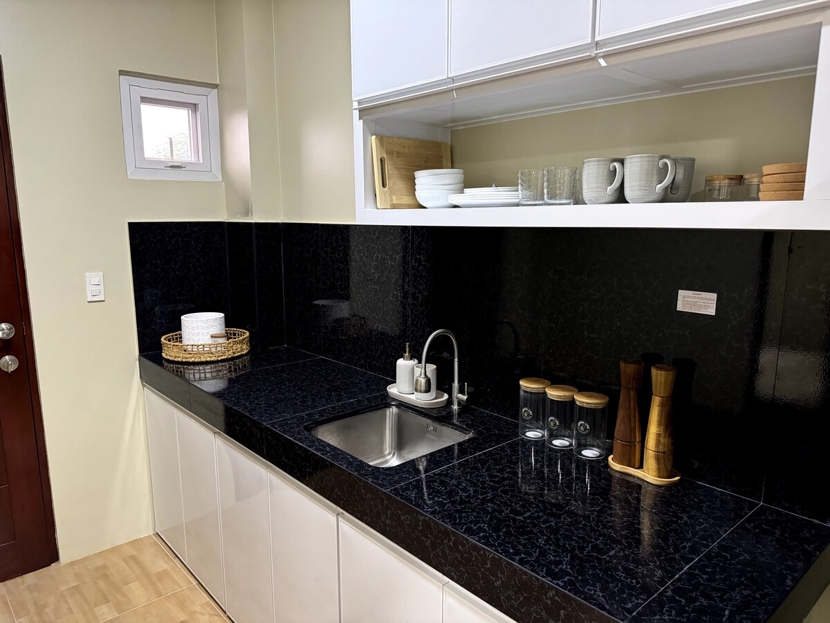 A modern kitchen area features sleek black countertops paired with glossy white cabinets. A sink is positioned centrally, with jars and kitchen utensils nearby, alongside neatly arranged glassware and tableware on the upper shelf. Natural light enters through a small window.