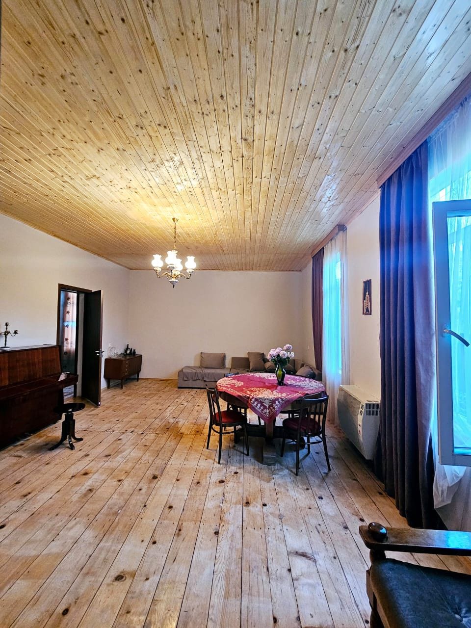 A spacious room features a wooden ceiling and warm-toned wooden floorboards. Natural light is provided by large windows, illuminating a table set for dining with a colorful tablecloth and fresh flowers. Comfortable seating is arranged in a cozy corner opposite a classic wooden sideboard.