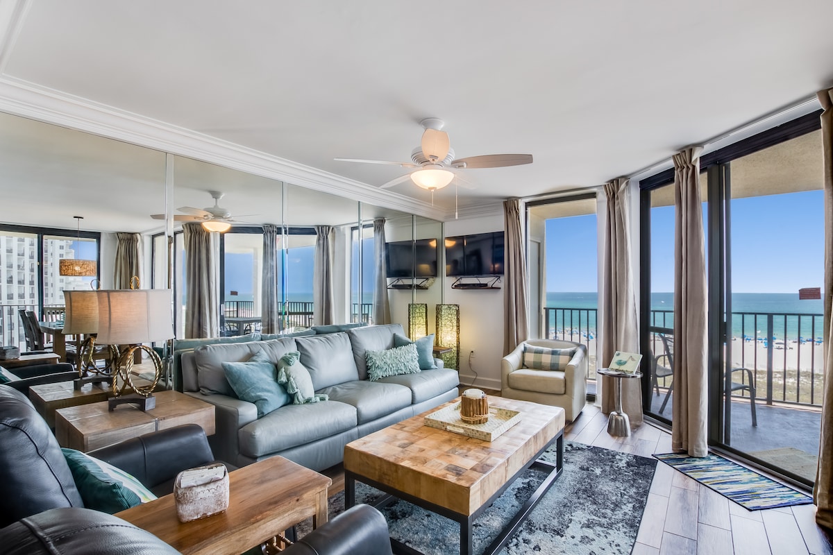 A spacious living area features comfortable seating and a wooden coffee table. Large glass doors provide unobstructed views of the beachfront, while light-colored curtains allow natural light to fill the room. A flat-screen TV is mounted on the wall, enhancing the modern decor.