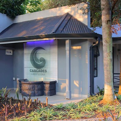 The entrance of the Cascades Guest Lodge is shown, featuring a contemporary sign with the lodge's logo. A stone feature wall is paired with a sleek, gray exterior. Lush greenery surrounds the area, enhancing the inviting atmosphere of the entrance.