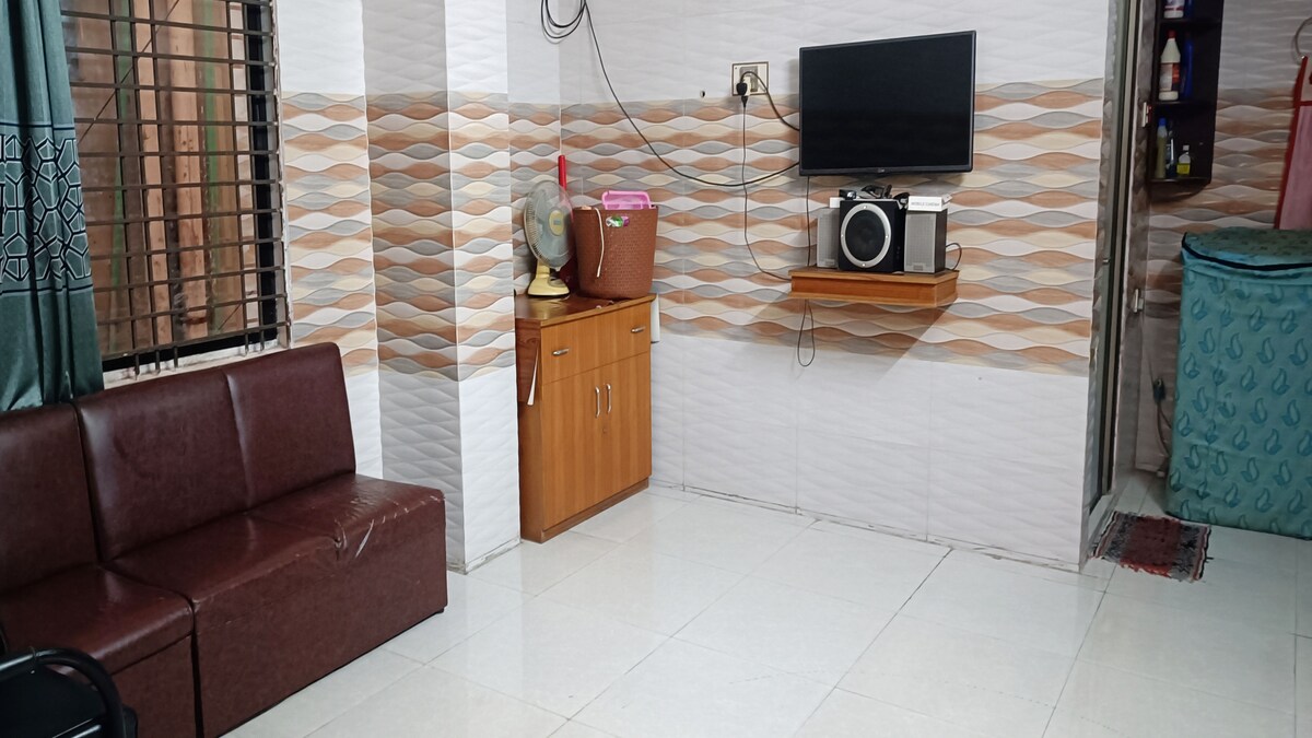 A cozy living area features a dark brown sofa and a wooden cabinet, equipped with a television and speakers. The walls display a modern wave-patterned design, and a fan is placed on a small table. Natural light enters through the window covered by sheer curtains.