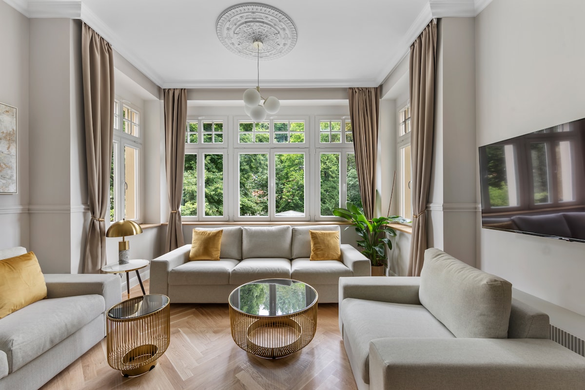 A well-appointed living area is presented with large windows that allow natural light to flood the space. Two sofas and a glass coffee table are arranged around a central area, complemented by elegant drapes and decorative elements, including a plant and a lamp.