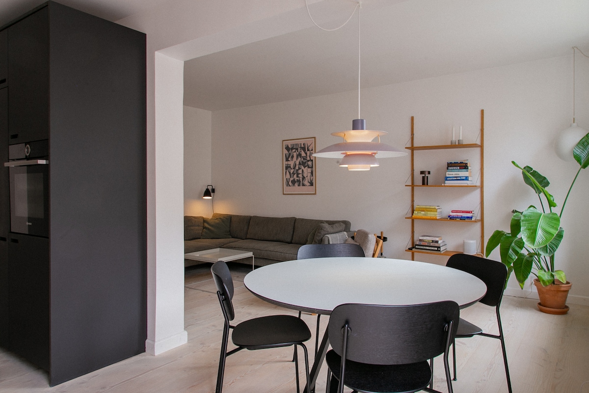 A dining area is displayed, featuring a round white table surrounded by six black chairs. A modern pendant light hangs above the table. In the background, a comfortable gray sofa is positioned against a wall, accompanied by a wooden bookshelf filled with books and a potted plant in a corner.