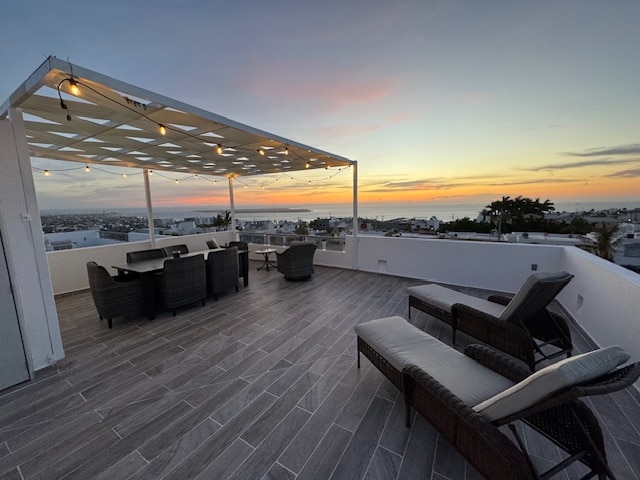 A spacious outdoor terrace is furnished with comfortable seating and lounge chairs. A pergola with string lights offers shade, and a panoramic view of the ocean and cityscape is visible as the sun sets, casting warm colors across the sky.