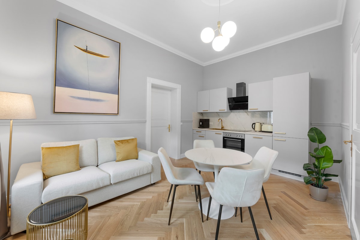 A modern kitchen and living area feature sleek cabinetry and a minimalist design. A circular dining table is surrounded by light-colored chairs, while a plush sofa with golden cushions provides seating. Natural light fills the space, highlighting the herringbone parquet flooring.