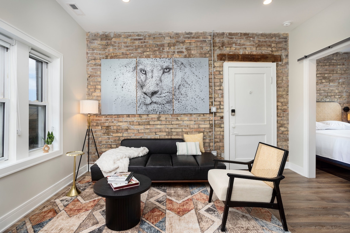 The living area features an exposed brick wall complemented by a modern black sofa and a patterned area rug. A round coffee table sits in front of the sofa, while a stylish chair provides additional seating. Artwork of a lion hangs prominently above the couch.