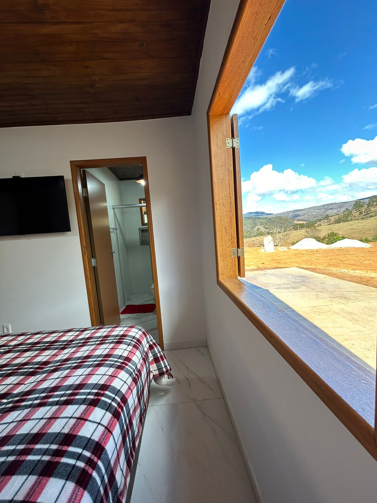 A spacious bedroom features a large window that opens to a scenic outdoor view. A flat-screen TV is mounted on the wall, and a checkered blanket covers the inviting bed. A wooden ceiling adds warmth to the room’s modern design.