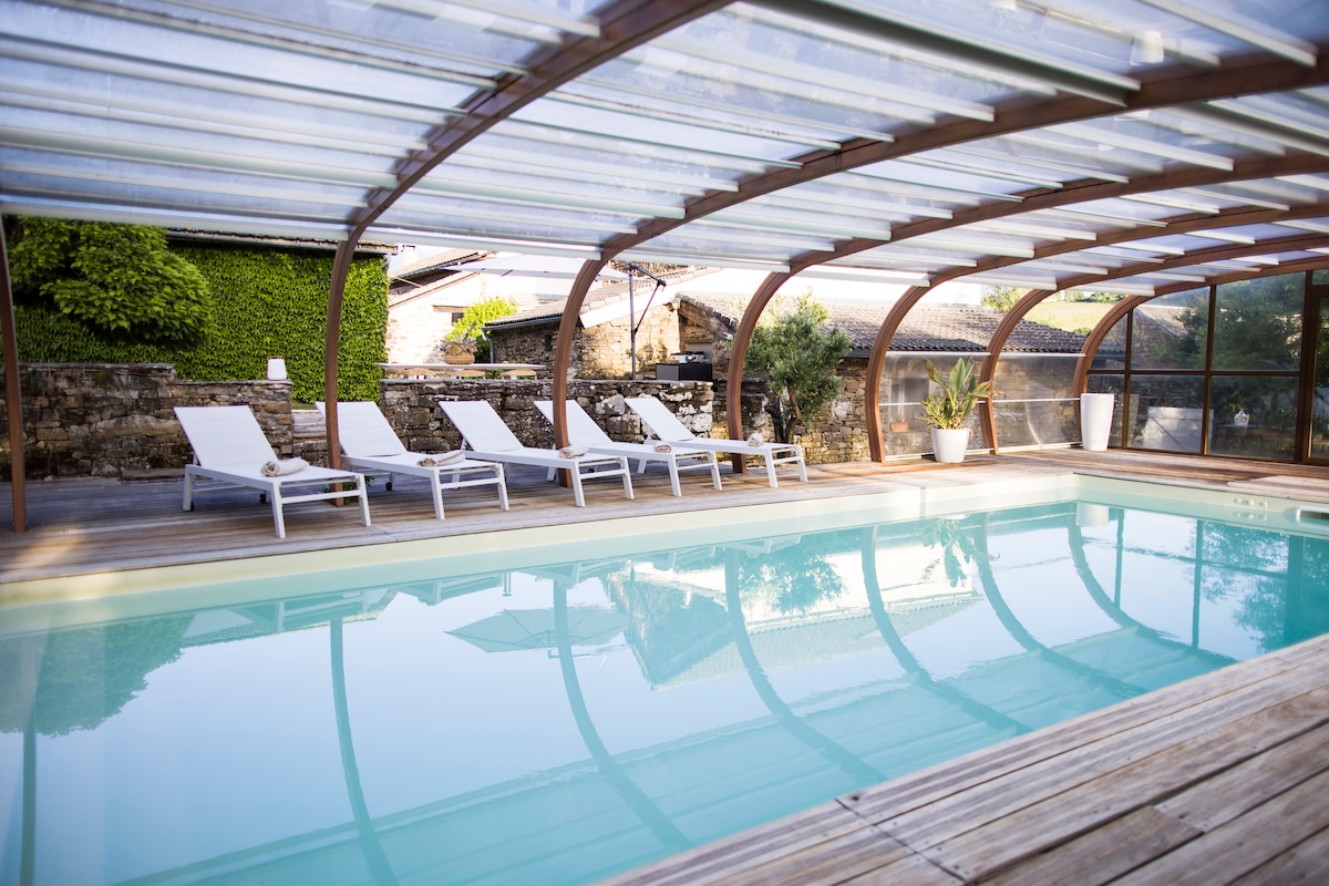 An indoor pool area features a clear cover allowing natural light to illuminate the space. Comfortable lounge chairs are arranged around the pool. Lush greenery is visible through the transparent walls, creating a serene atmosphere. Warm wooden accents enhance the overall design.