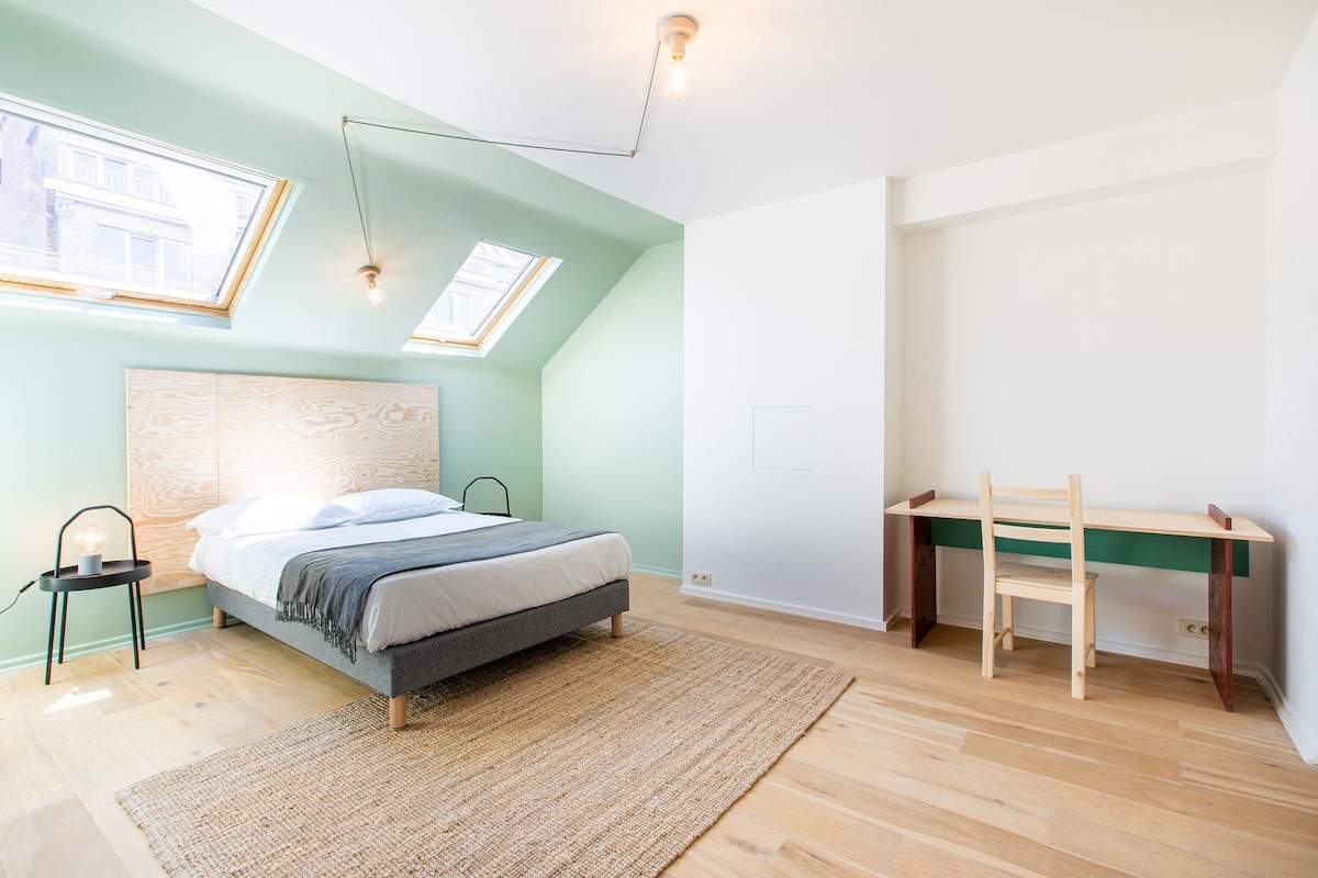 A simple bedroom features a spacious bed with crisp white linens, positioned beneath skylights. A minimalist desk with a chair is situated against a green accent wall, while natural light brightens the room. A woven rug complements the wooden flooring.