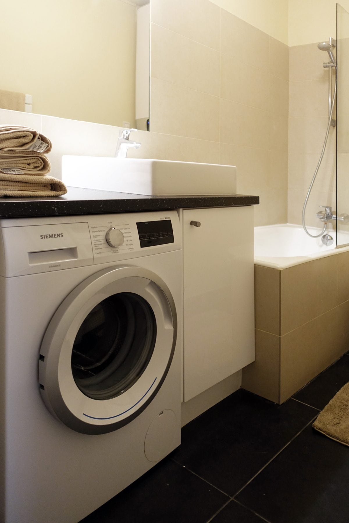 A functional bathroom area includes a washing machine and a compact sink, accompanied by neatly stacked towels. A shower is visible alongside a bathtub, with neutral-colored tiles adorning the walls and floor, enhancing the space's practicality.