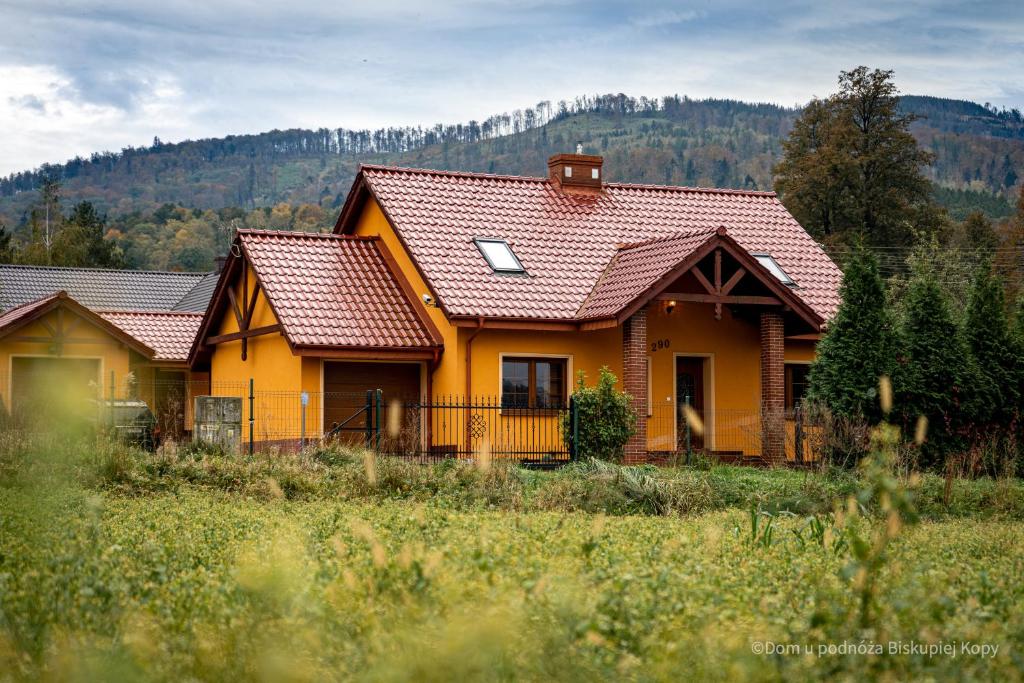 A yellow house with a red tiled roof is surrounded by greenery, featuring a spacious front yard. The structure has a prominent gabled entryway and a chimney. Lush trees and rolling hills serve as a backdrop, showcasing the picturesque mountain landscape.