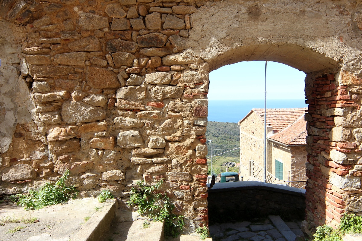 An arched stone doorway frames a view of the coastline, revealing distant blue waters and hills. Sunlight casts gentle shadows across the rough stone walls, while greenery peeks through at the base, hinting at the surrounding natural beauty.