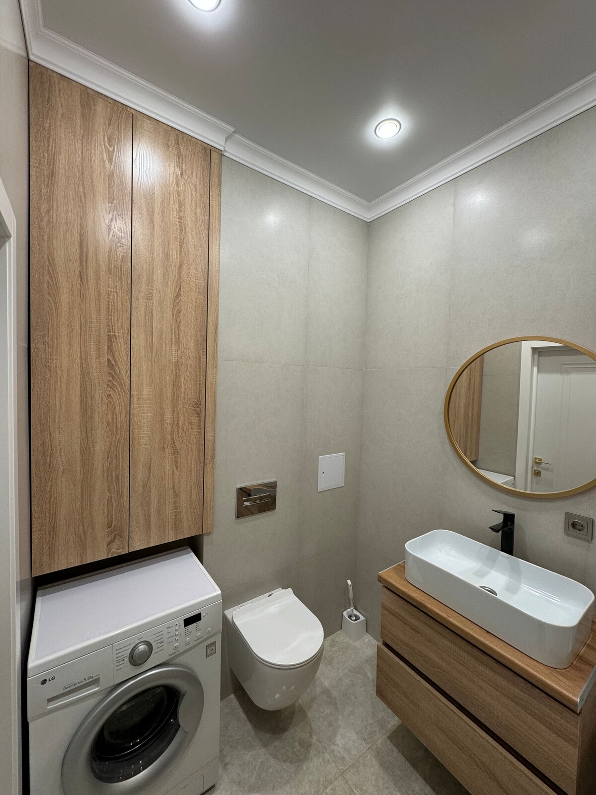 A modern bathroom features light gray walls and wood cabinetry. A round mirror reflects a well-lit space above a sleek basin. A compact washing machine is incorporated into the cabinetry, and a comfortable toilet is positioned nearby, contributing to the functional layout.