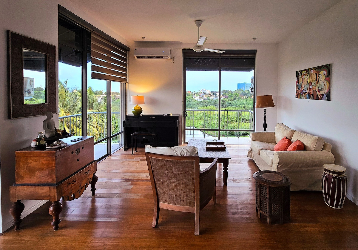 A spacious living area features large glass doors that open to a balcony, allowing views of greenery. Comfortable seating options include a cream-colored sofa and a woven chair. A wooden side table and a decorative drum add character to the space, complemented by warm lighting.