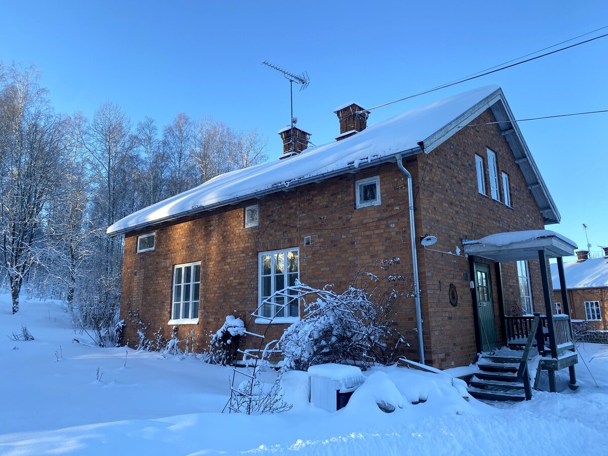 The exterior of a charming brick house is shown, resting under a blanket of fresh snow. Frost-covered trees surround the property, complementing the serene winter landscape. The front porch features steps leading to a green door, enhancing the inviting look of the home.