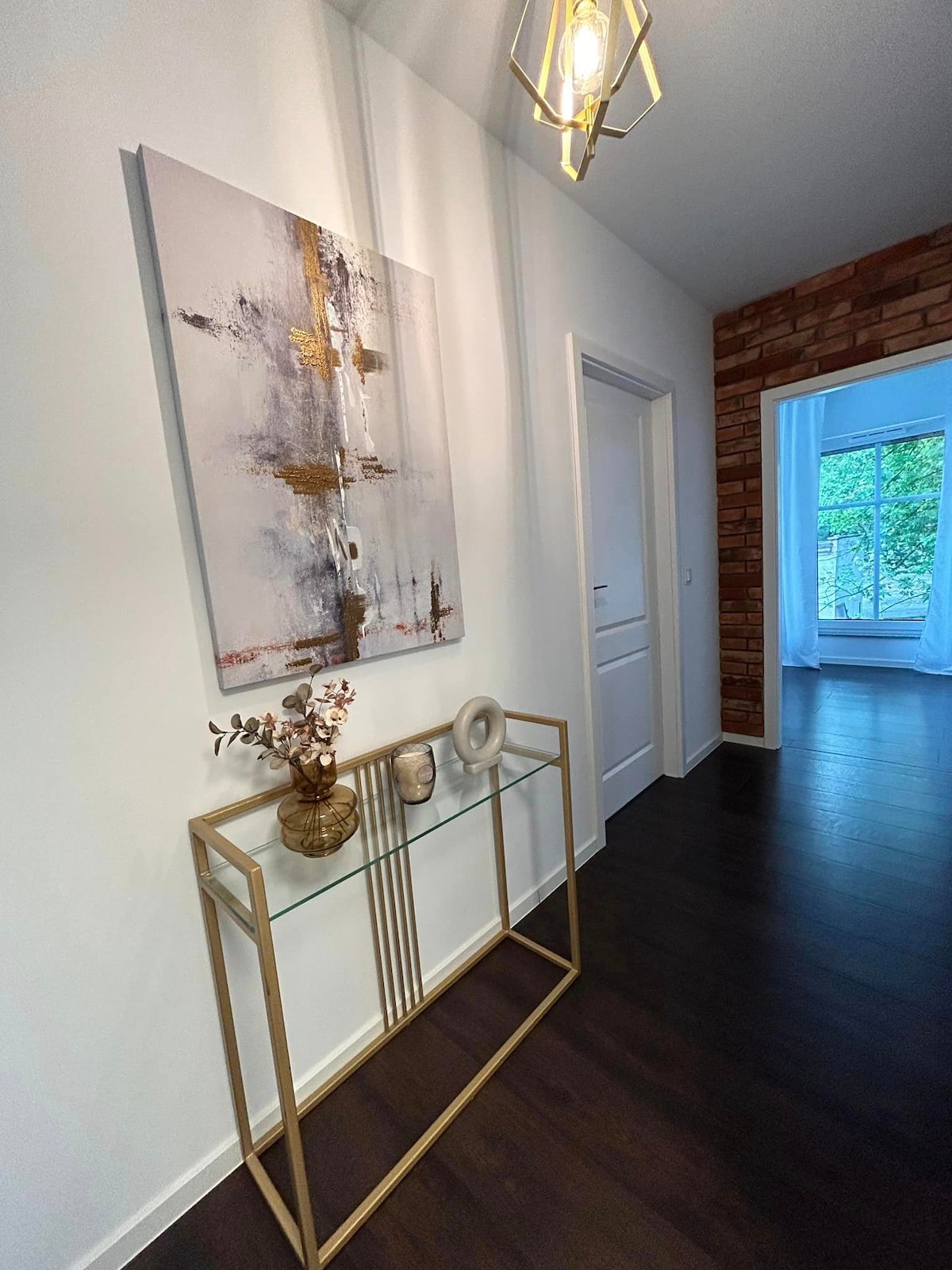 A narrow hallway displays a modern piece of art with neutral colors and gold accents on a white wall. A decorative console table, featuring a glass top and gold frame, holds a vase and a sculptural object. Natural light filters in from a nearby room.