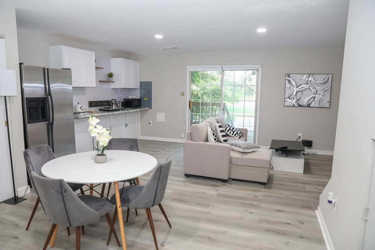An open living space features a light-filled area with a round dining table surrounded by four grey chairs. A comfortable sofa is positioned beside a coffee table, facing sliding glass doors that offer views of a green outdoor space.
