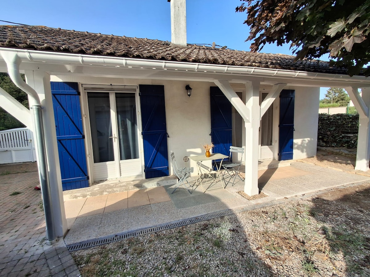 The exterior of the house features white walls and a tiled roof, complemented by blue shutters. A small outdoor seating area includes a table and two chairs, positioned on a stone patio. Natural light illuminates the space, with greenery in the background.