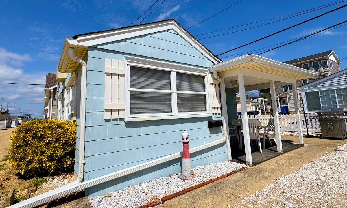 The exterior of a charming blue cottage is pictured, featuring white shutters and a covered entrance. A beige path leads to the porch, where a small table and chairs are visible. White gravel decorates the garden area alongside neatly trimmed yellow shrubbery.